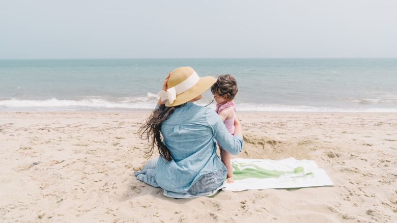 A woman with a hat sits on the beach, holding a child, both facing the ocean on a sunny day, with a towel beneath them in the sand.