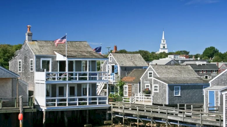 The image shows a quaint seaside village with wooden houses, American flags, a pier, and a white church tower in the background.