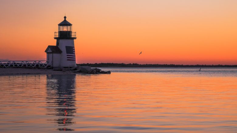 A lighthouse with an American flag at sunset stands on a calm water's edge, reflecting colors of the sky, with a single bird flying nearby.