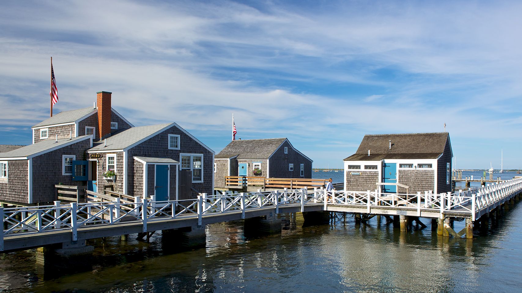 Harbor view with wooden buildings on a pier, surrounded by water and boats, under a blue sky with scattered clouds.