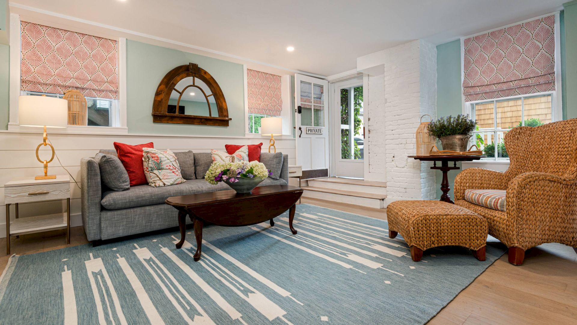 A cozy living room with a gray sofa, wicker chair, blue-striped rug, round mirror, side lamps, and pink window treatments.
