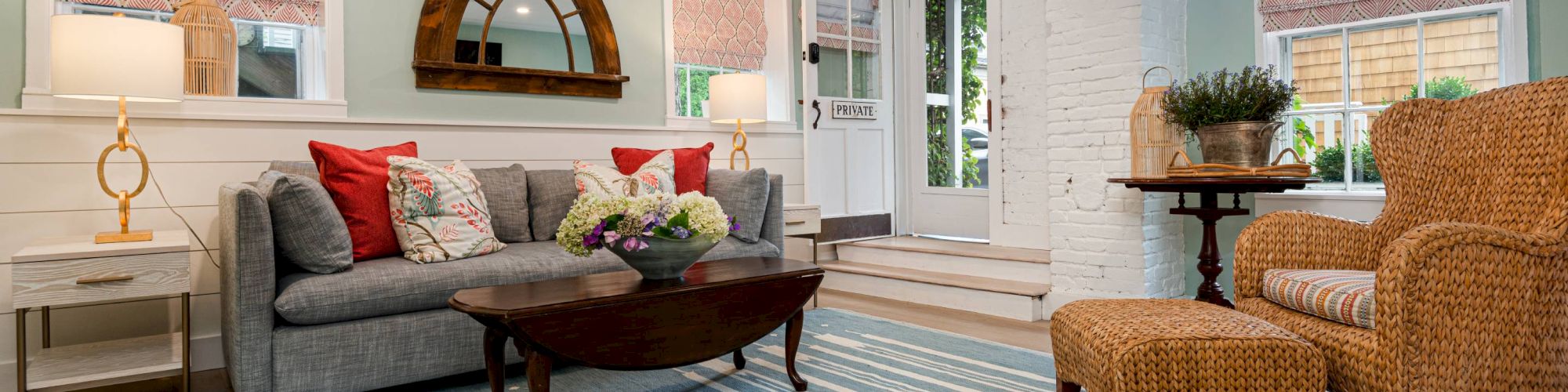 A cozy living room with a gray sofa, wicker chair, blue-striped rug, round mirror, side lamps, and pink window treatments.