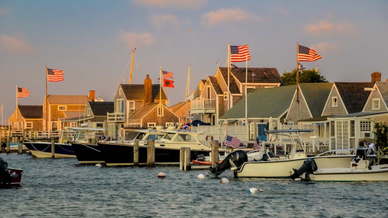 The image shows a coastal scene with boats docked near wooden houses flying multiple American flags, captured during sunset on a calm day.