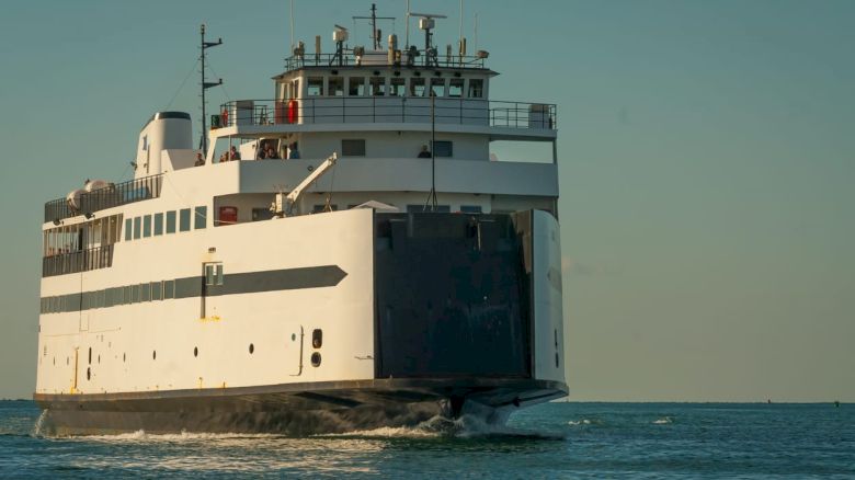 The image shows a large ferry navigating through the water, with a clear sky in the background and passengers visible on the deck.