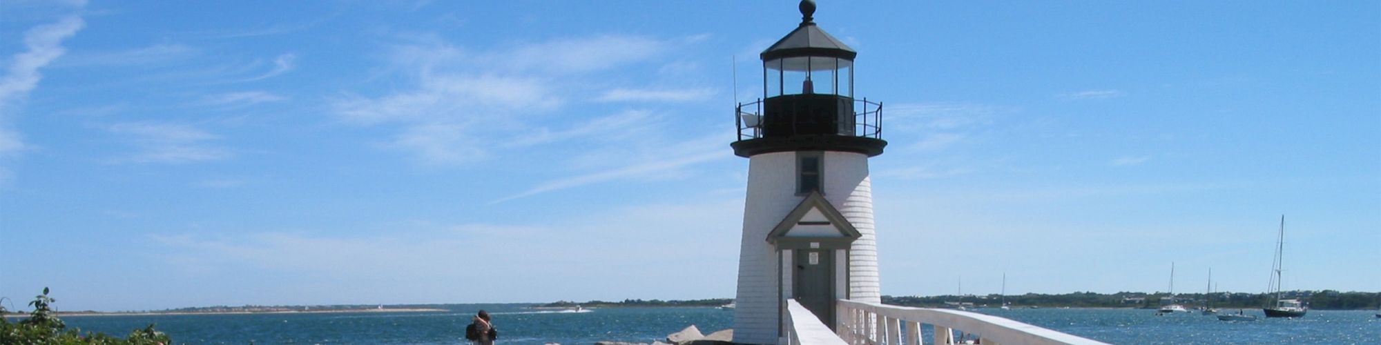 A sandy beach with a lighthouse and a white fence along a wooden path leading towards it under a bright blue sky with some scattered clouds.