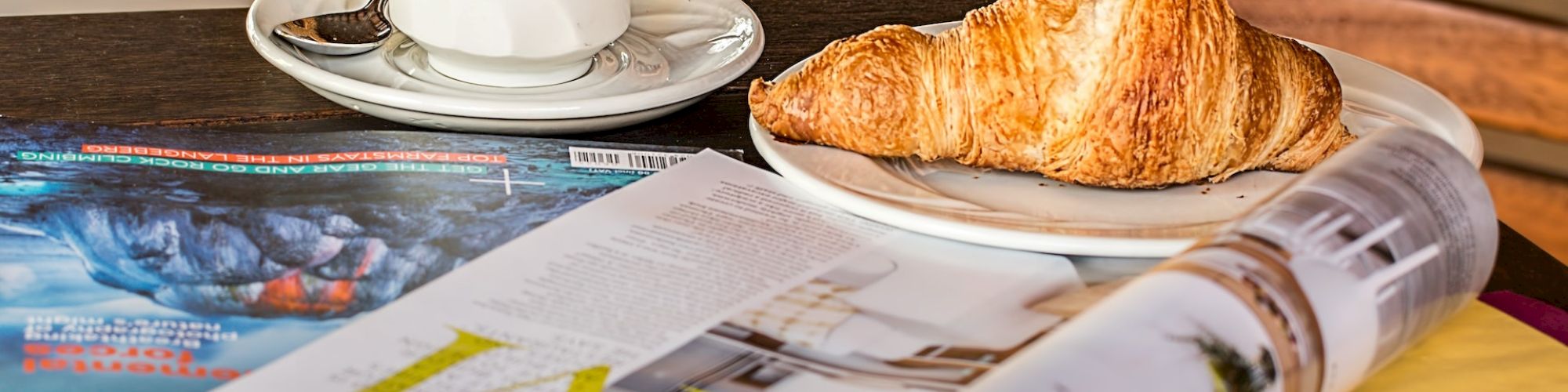 A cup of coffee, a croissant on a plate, and an open magazine are placed on a wooden table with a chair in the background.