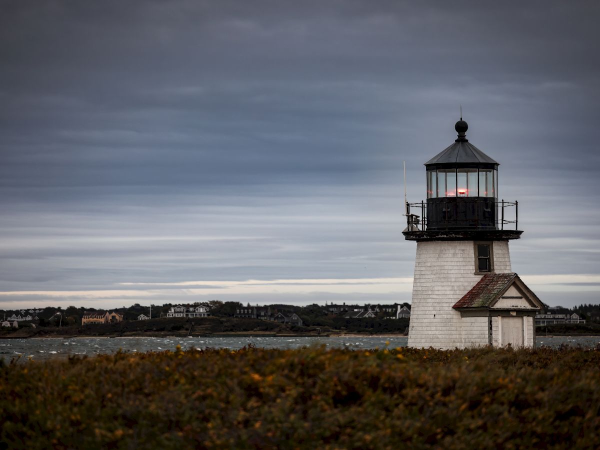 The image shows a white lighthouse with a dark roof on a grassy coastline under a cloudy sky, with distant buildings visible across the water.