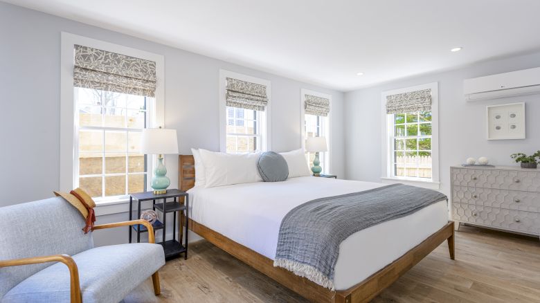 A bright bedroom with a large bed, gray chair, two bedside tables with lamps, and a dresser, featuring three windows and wooden floors.