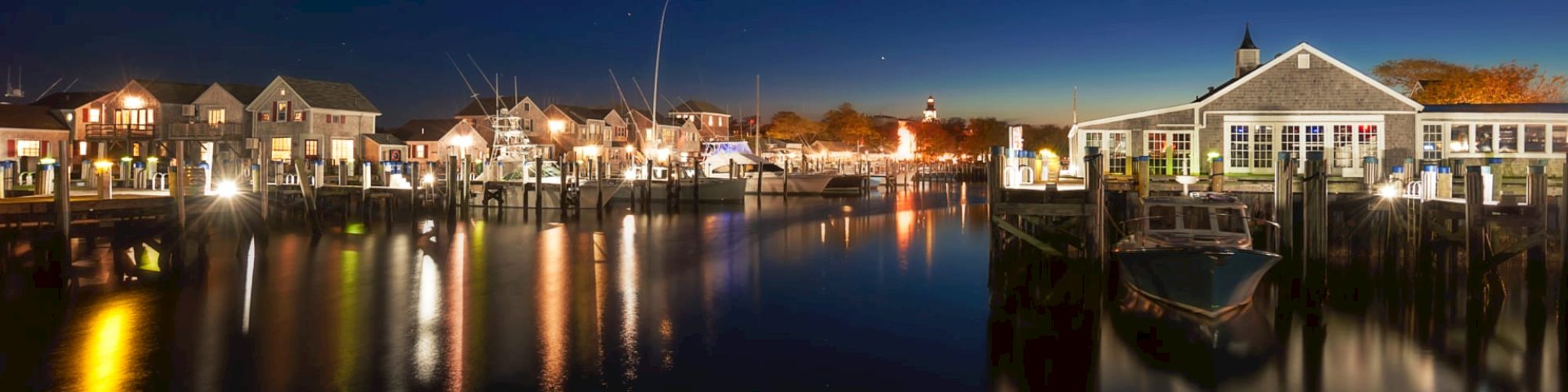 A nighttime waterfront scene features illuminated buildings reflecting on calm water, boats docked, and a lighthouse visible in the distance.