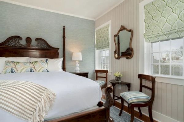 A cozy bedroom with a large bed, wooden furniture, mirrors, two chairs, a side table, a lamp, and windows with green Roman shades.