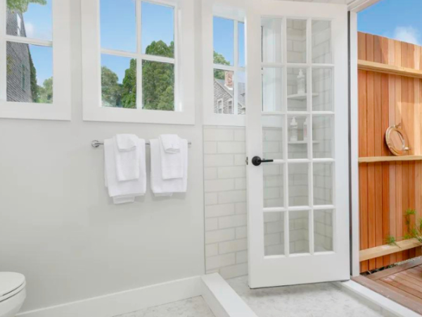 A bright bathroom with white towels, large windows, a glass door leading to an outdoor wooden deck with plants and decor.
