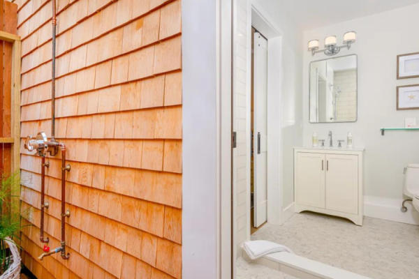 This image shows an outdoor shower next to an indoor bathroom with a toilet, vanity, and mirror. The bathroom is white with modern fixtures.