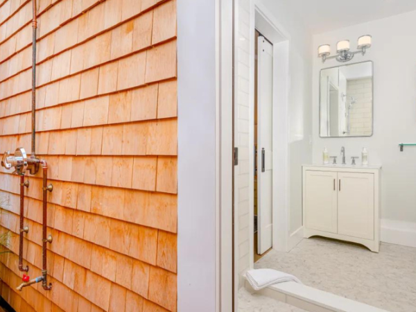 This image shows an outdoor shower next to an indoor bathroom with a toilet, vanity, and mirror. The bathroom is white with modern fixtures.