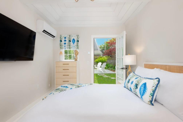 A cozy bedroom features a wall-mounted TV, air conditioner, dresser, and a bed with white linens and blue accents, leading to a garden view.