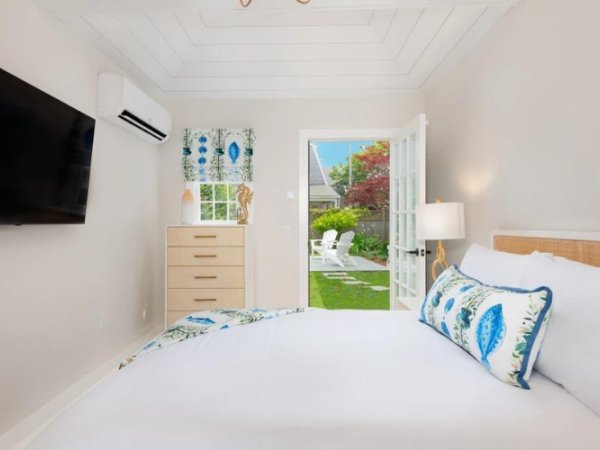 A cozy bedroom features a wall-mounted TV, air conditioner, dresser, and a bed with white linens and blue accents, leading to a garden view.