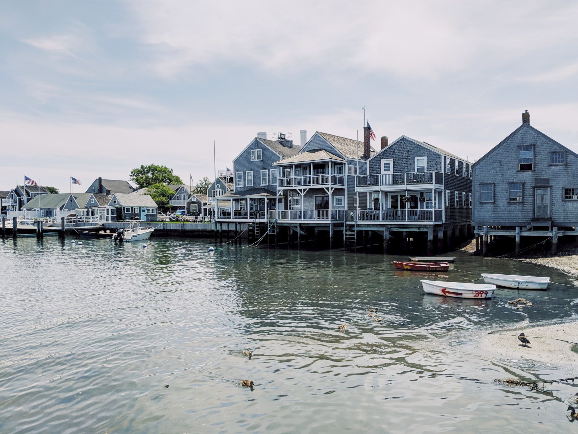 The image shows a serene coastal scene with several wooden houses built on stilts over the water, and small boats floating nearby.