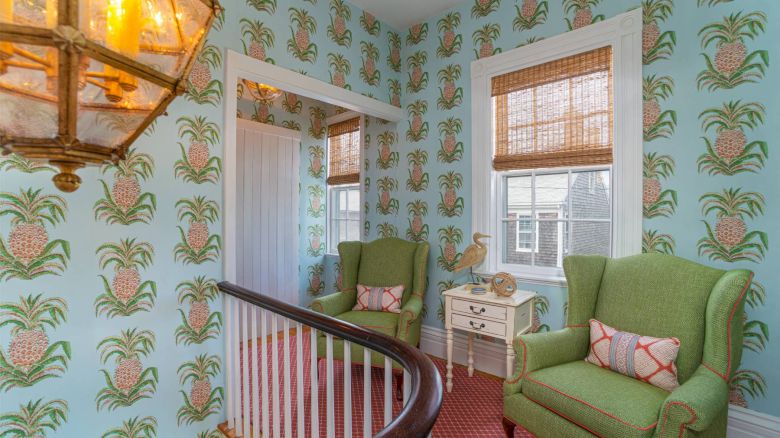 A room with pineapple-patterned wallpaper, green armchairs, a small white table, a lantern-style light fixture, and windows with bamboo blinds.