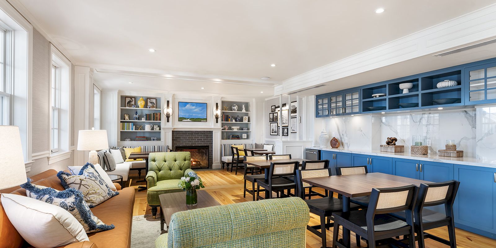 A cozy living room with a fireplace, sofas, bookshelves, blue kitchen cabinets, and dining tables, all in a bright, open space.