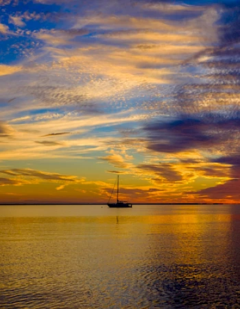A boat is silhouetted against a vibrant sunset over calm waters, with dramatic clouds painted in various shades of orange, blue, and purple.