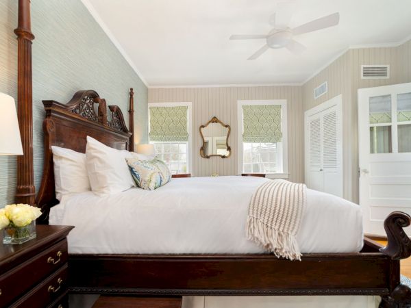 A bright traditional bedroom with a wooden sleigh bed, white linens, patterned pillows, dresser, mirror, and ceiling fan.