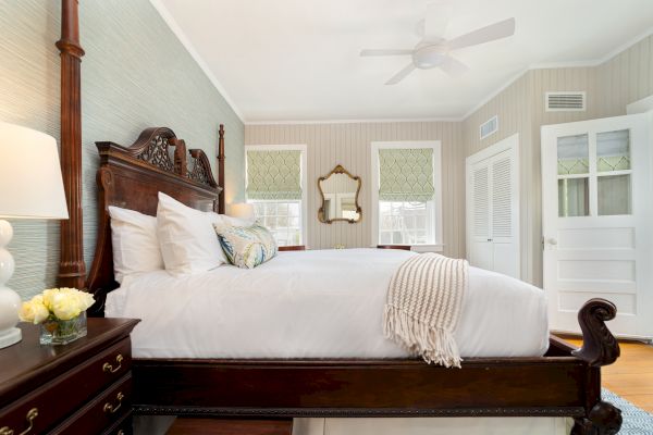 A cozy bedroom with a wooden bed, white bedding, a nightstand with flowers, a mirror, and windows with blinds. All walls are painted in white.