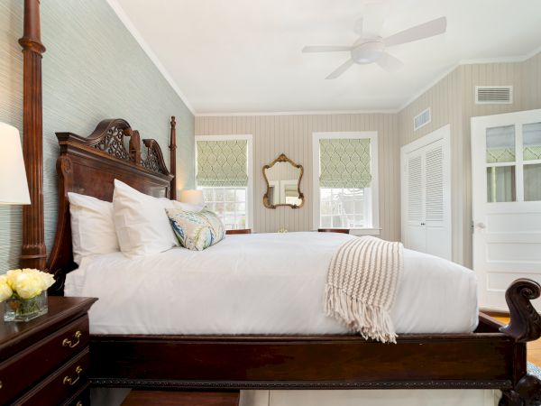 A cozy bedroom with a wooden bed, white bedding, a nightstand with flowers, a mirror, and windows with blinds. All walls are painted in white.