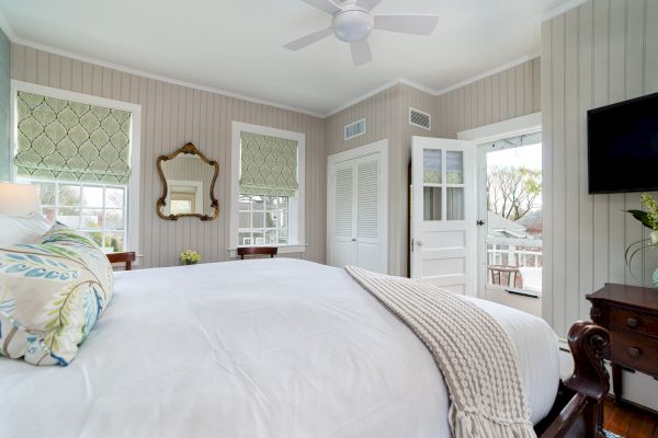 A cozy bedroom with a large bed, decorative pillows, a blanket, a wall mirror, a ceiling fan, a TV, and a door opening to a porch with outside views.