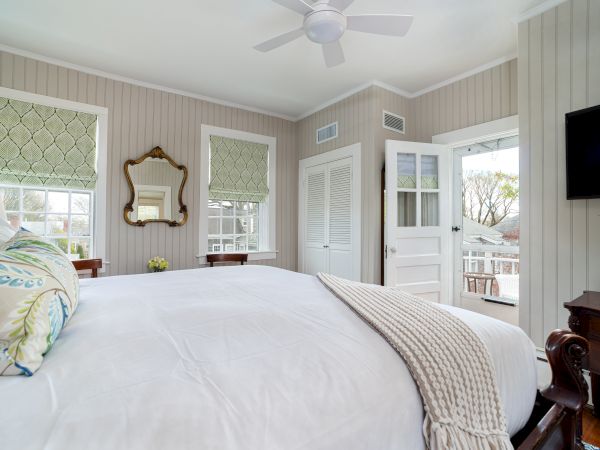 A cozy bedroom with a large bed, decorative pillows, a blanket, a wall mirror, a ceiling fan, a TV, and a door opening to a porch with outside views.