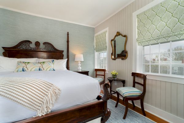 A cozy bedroom with an ornate wooden bed, white linens, two striped chairs, a lamp, mirrors, and large windows with patterned shades.