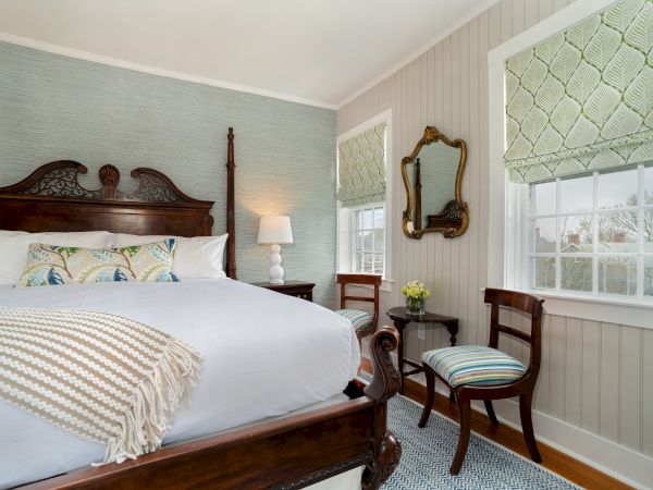 A cozy bedroom with an ornate wooden bed, white linens, two striped chairs, a lamp, mirrors, and large windows with patterned shades.