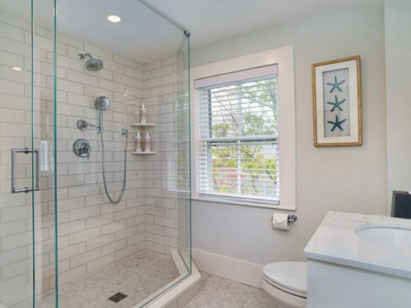 Bright bathroom with a glass shower, tiled walls, toilet, vanity sink, window, and framed wall art.