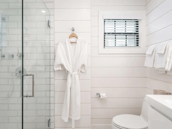 A modern bathroom with a glass shower, white bathrobe, toilet, towels, and a small window with white blinds, all set in a white-tiled space.