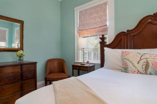 A cozy bedroom with a wooden bed, floral pillows, dresser with mirror, chair, side table with lamp, and window with blinds.