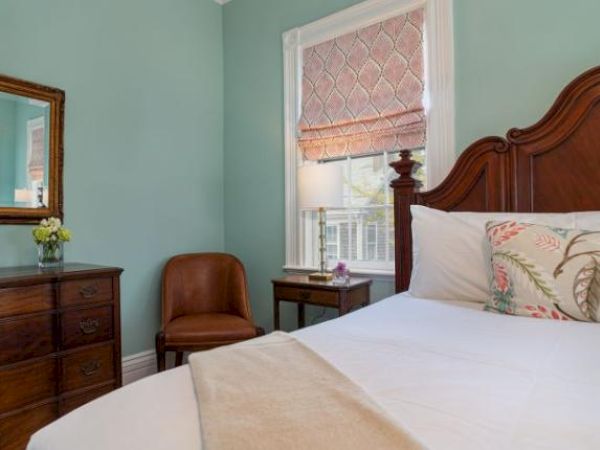 A cozy bedroom with a wooden bed, floral pillows, dresser with mirror, chair, side table with lamp, and window with blinds.
