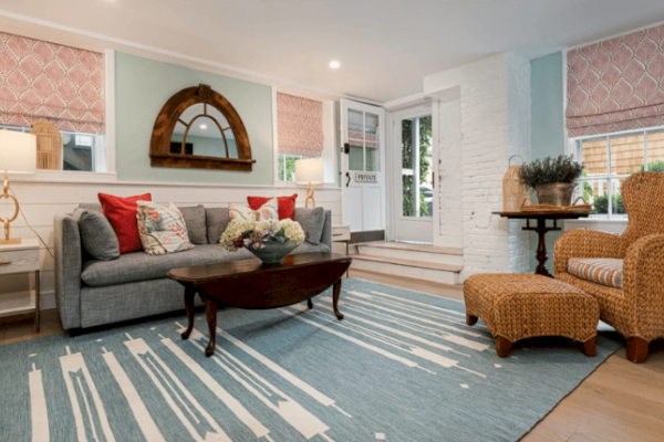 A cozy living room with a gray couch, red and white pillows, wicker chair and ottoman, wooden coffee table, and blue rug. Lamp and decor included.