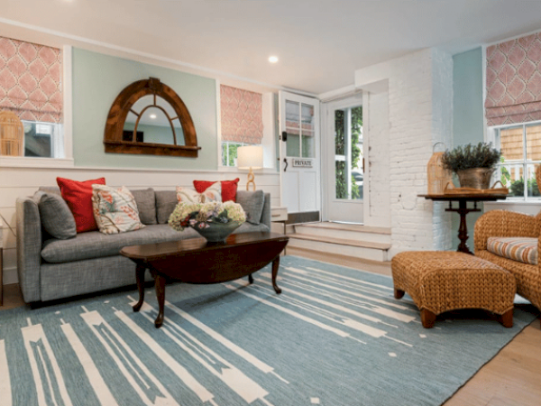 A cozy living room with a gray couch, red and white pillows, wicker chair and ottoman, wooden coffee table, and blue rug. Lamp and decor included.