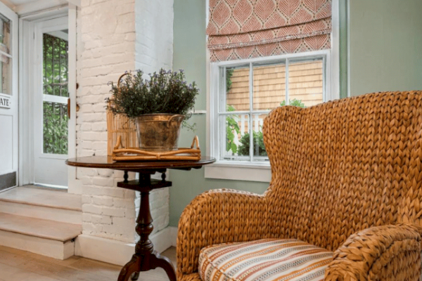 A cozy seating area with a wicker armchair, a round table holding a plant and book, white walls, and a window showcasing a sunny outdoor view.