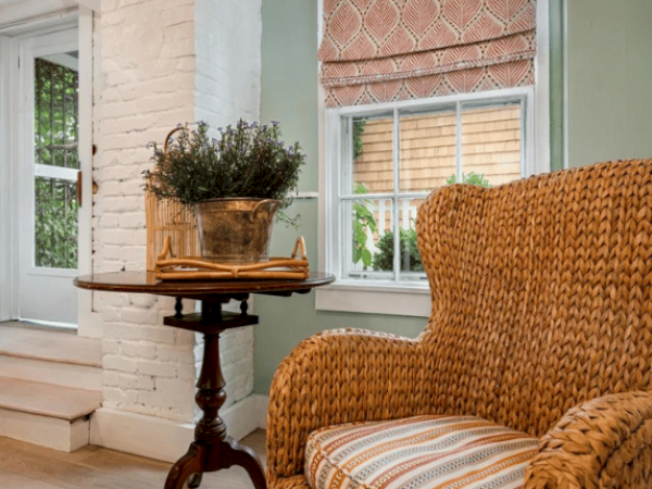 A cozy seating area with a wicker armchair, a round table holding a plant and book, white walls, and a window showcasing a sunny outdoor view.