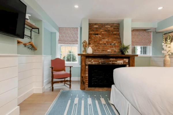 A cozy bedroom with a brick fireplace, a mounted TV, a plush armchair, a bed, and a decorative rug underfoot.