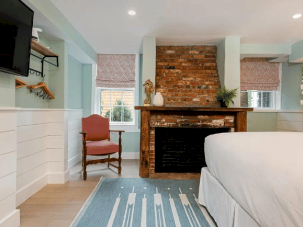 A cozy bedroom with a brick fireplace, a mounted TV, a plush armchair, a bed, and a decorative rug underfoot.