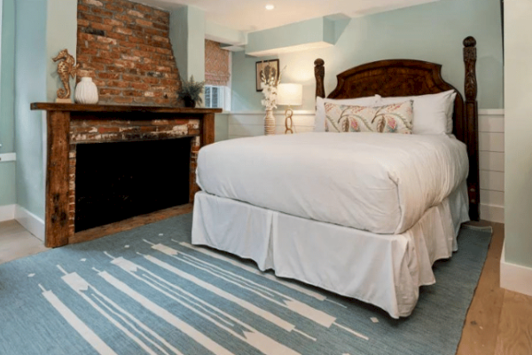 A cozy bedroom with a white bed, brick fireplace, decorative mantle, and a blue patterned rug on wooden floors. Light blue walls and soft lighting.