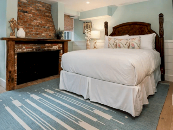 A cozy bedroom with a white bed, brick fireplace, decorative mantle, and a blue patterned rug on wooden floors. Light blue walls and soft lighting.