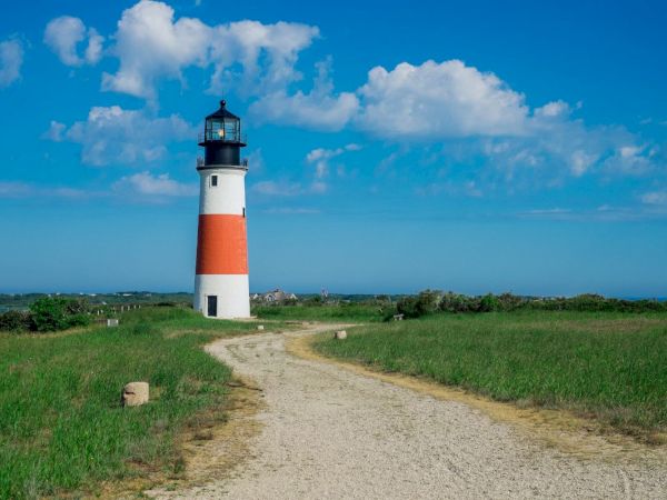 A white-and-orange lighthouse stands by a dirt path under a vivid blue sky with big fluffy clouds.