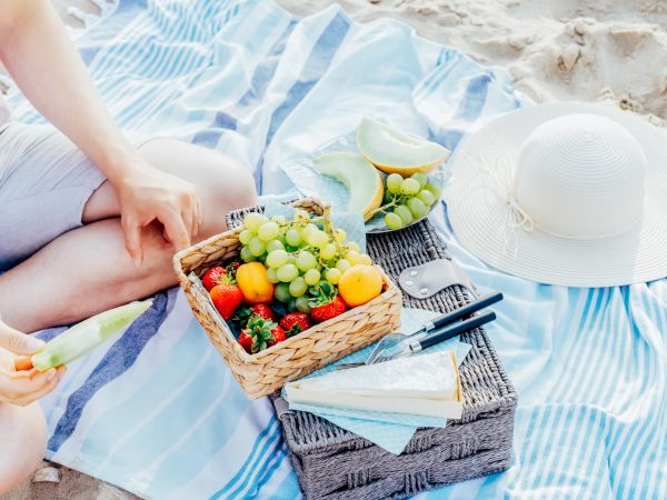 A beach picnic scene with a person, a basket of fruits, a hat, and a striped mat. The basket contains grapes, strawberries, and other fruits.