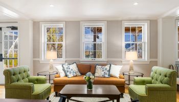 A bright living room with a tan sofa, two green armchairs, blue-pattern pillows, and three windows.