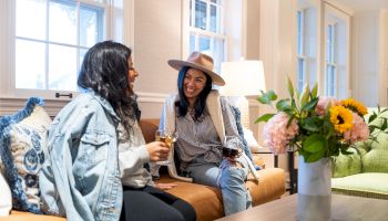 Two people sitting on a couch, enjoying drinks and chatting. A vase with flowers is on the table. The room has bright, natural light.