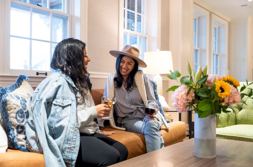 Two people sitting on a couch, enjoying drinks and chatting. A vase with flowers is on the table. The room has bright, natural light.