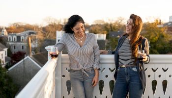 Two people are standing on a balcony, smiling and holding drinks, with a scenic view of houses and trees in the background.