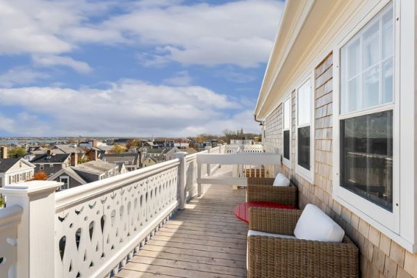 A balcony with a wooden deck, wicker furniture, and a white railing overlooking a town under a cloudy sky.