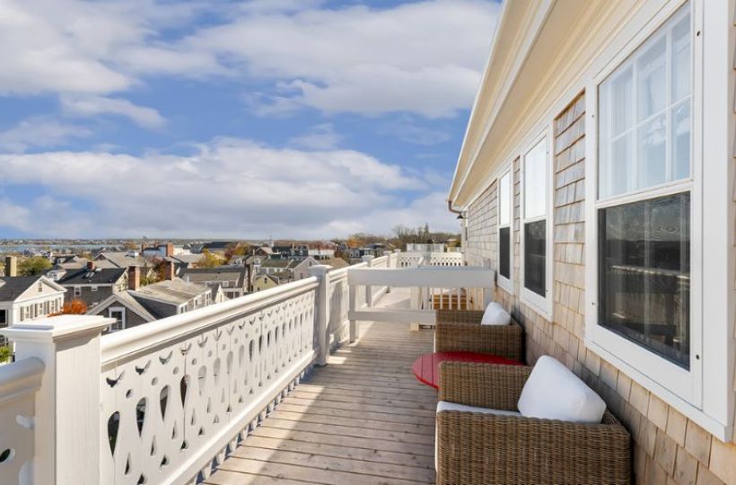 A rooftop terrace with wicker chairs and cushions, offering a view of nearby rooftops and a distant sea under a partly cloudy sky.